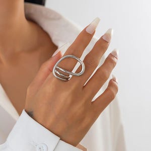 Op de afbeelding: Een zilverkleurige ring met een uniek, abstract ontwerp. De ring heeft een grote ovale vorm met twee parallelle banden en een gebogen lijn, wat een moderne en artistieke uitstraling creëert. De ring wordt aan een vinger gedragen.