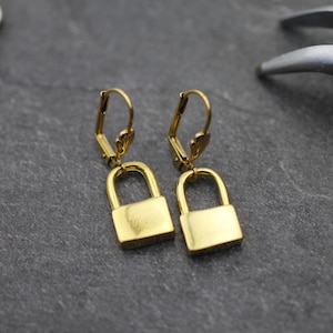 May include: A pair of gold-coloured padlock earrings. Each earring has a lever back closure and a rectangular padlock charm. The earrings are displayed on a dark grey surface.