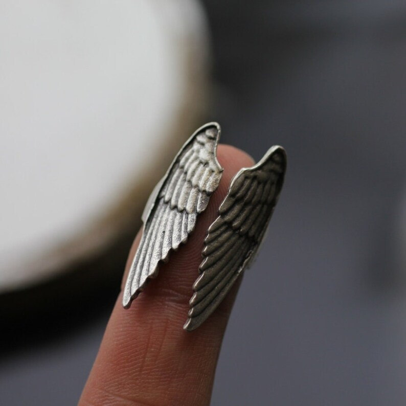 May include: A silver-colored ring shaped like angel wings. The ring is designed to wrap around a finger, with each wing extending on either side. The wings have detailed feather textures, creating a realistic appearance. The ring is shown on a finger.