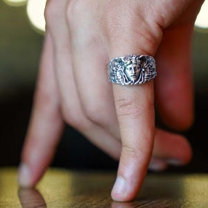 Puede incluir: Un anillo plateado con un diseño detallado de la cara de Medusa. El anillo se lleva en un dedo, mostrando las intrincadas tallas y texturas del metal. El diseño del anillo incluye una cara central rodeada de cabello fluido y detalles ornamentados.