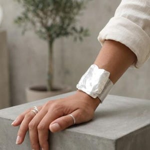 May include: A wide, silver-toned cuff bracelet with a textured, hammered surface finish. The bracelet is worn on the wrist, accompanied by two thin, plain silver-toned rings on the fingers, showcasing a metallic sheen and irregular, organic contours.