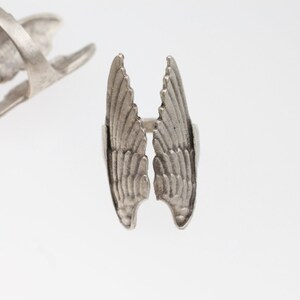 May include: A silver-toned ring featuring a detailed wing design. The ring's wings are spread open, showcasing intricate feather detailing. Another ring with a similar design is visible in the background, set against a white backdrop.