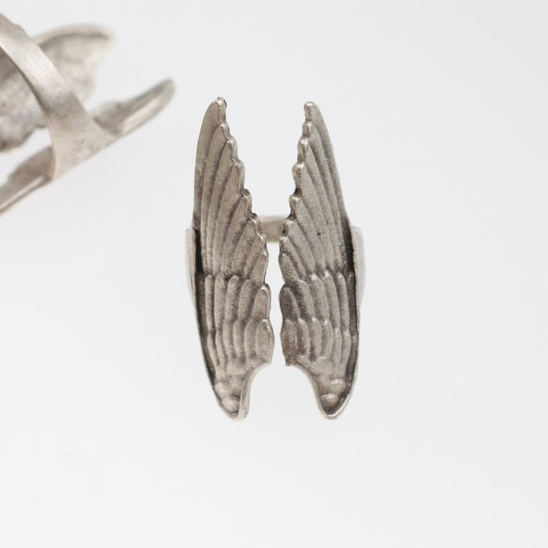May include: A silver-toned ring featuring a detailed wing design. The ring's wings are spread open, showcasing intricate feather detailing. Another ring with a similar design is visible in the background, set against a white backdrop.