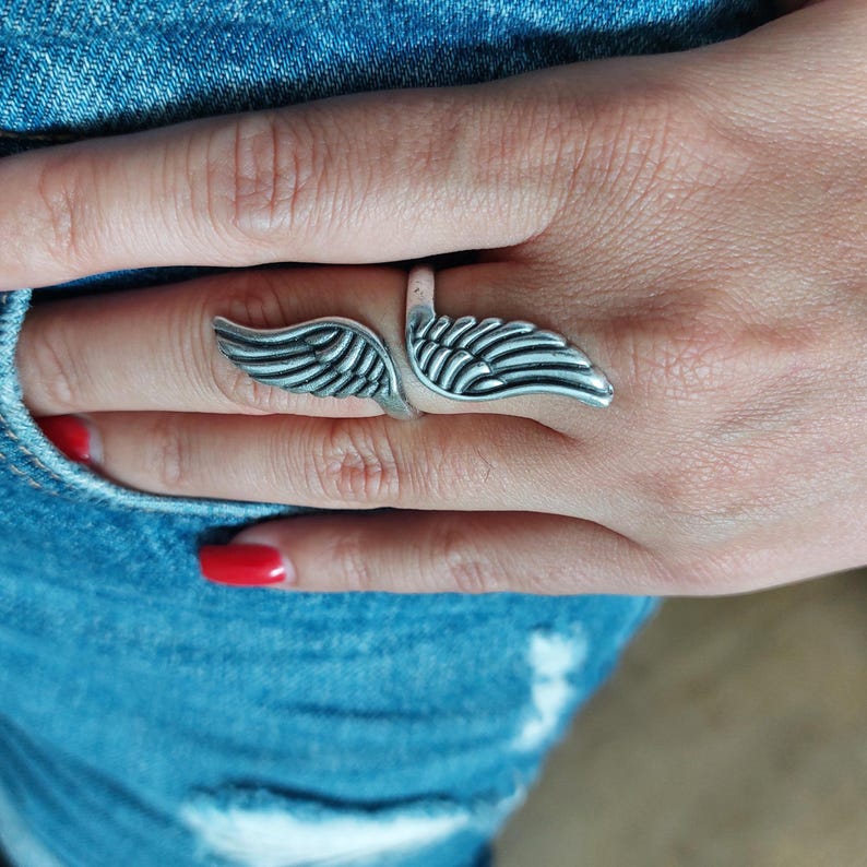 K&ouml;nnte beinhalten: Ein silberfarbener Ring mit offenem Design, der zwei stilisierte Fl&uuml;gelformen aufweist. Der Ring wird an einem Finger getragen, wobei sich die Fl&uuml;gel nach au&szlig;en erstrecken. Das Design des Rings ist detailliert, mit Linien, die Federn andeuten.