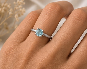 May include: A silver-toned ring featuring a single round-cut light blue gemstone held in a four-prong setting. The band is smooth and polished, showcasing a minimalist design that highlights the central stone's pale blue color and clarity.