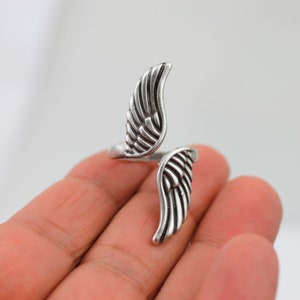 K&ouml;nnte beinhalten: Ein silberfarbener Ring mit detailliertem Fl&uuml;geldesign. Der Ring umschlie&szlig;t den Finger mit zwei stilisierten Fl&uuml;geln, die in entgegengesetzte Richtungen weisen. Die Fl&uuml;gel haben strukturierte Federdetails, was ein zartes Accessoire ergibt.