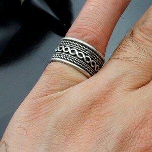 Może przedstawiać: Srebrny pierścionek z plecionym wzorem. Pierścionek jest noszony na palcu.