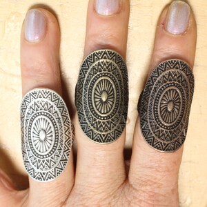 Könnte beinhalten: Drei ovale Ringe in Silber-, Bronze- und Messingtönen. Jeder Ring hat ein kompliziertes, symmetrisches Muster mit einem zentralen Design und geometrischen Details. Die Ringe werden auf Fingern präsentiert.