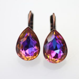 Puede incluir: Un par de pendientes en forma de lágrima con un acabado de cobre. Los pendientes presentan cristales iridiscentes de color morado y naranja.