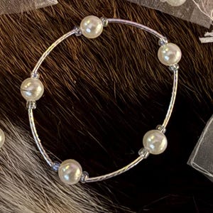 May include: A delicate bracelet featuring six faux pearls strung on a thin, silver-colored wire. The pearls are evenly spaced around the bracelet's circumference. The background is a dark, textured surface.