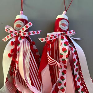 Peut inclure: Deux ornements décoratifs en forme de petits pots de confiture de fraises. Chaque pot rouge est surmonté d'un couvercle blanc et d'une étiquette indiquant "Homemade Strawberry Jam". Des rubans rouges, blancs et roses, ornés de fraises et de pompons, pendent des pots.