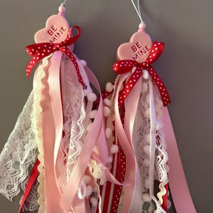 Könnte beinhalten: Zwei Valentinstagsornamente. Jedes Ornament hat einen herzförmigen Anhänger mit dem Text "BE MINE", eine rote Schleife mit Tupfen und fließende Bänder in Rosa, Weiß und Rot. Weiße Spitze und kleine weiße Pompons verleihen dem Design Textur.