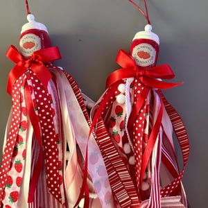 Peut inclure: Deux ornements décoratifs en forme de pots de confiture de fraises miniatures. Chaque pot est orné d'un nœud rouge et d'une cascade de rubans rouges, blancs et roses à motifs de fraises et à pois. Des pompons blancs sont attachés aux rubans.