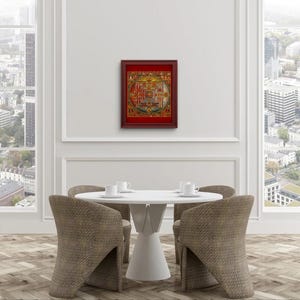 Op de afbeelding: Een ingelijst mandala-print met ingewikkelde details en levendige kleuren, hangend aan een witte muur boven een witte tafel met vier stoelen.