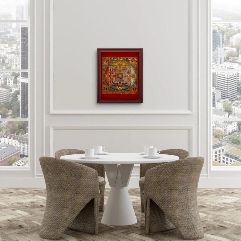 Op de afbeelding: Een ingelijst mandala-print met ingewikkelde details en levendige kleuren, hangend aan een witte muur boven een witte tafel met vier stoelen.