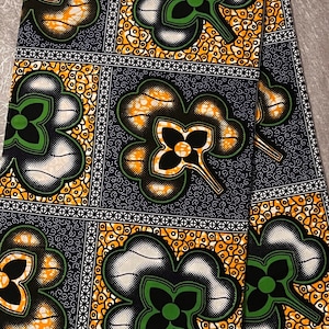 May include: A close-up of a blue and white fabric with a repeating pattern of green, black, and orange clover-shaped designs. The fabric has a textured surface and is labeled "BLOCK PRINTS HITAGET 29229".