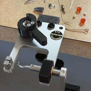 May include: A silver and black jewelry-making tool with the text "EYE PROTECTION REQUIRED". Various small metal and glass components, including beads, wires, and clasps, are scattered around the tool on a light brown surface. The tool has a curved arm and a black handle.