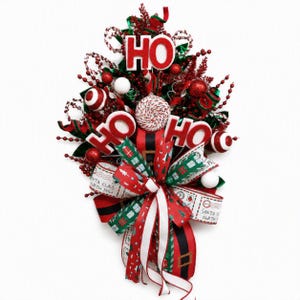 May include: A Christmas teardrop swag decoration featuring red and white HO HO HO lettering, a central peppermint candy ornament, red berry sprays, and green foliage. The base includes ribbons with Santa Claus North Pole postal stamps.