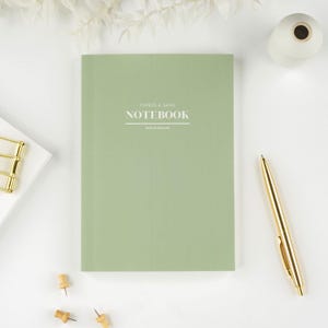 May include: A sage green notebook with the text "PAPERS & GEMS NOTEBOOK" and "MADE IN ENGLAND" on the cover. A gold pen, white paper, and push pins are arranged on a white surface.