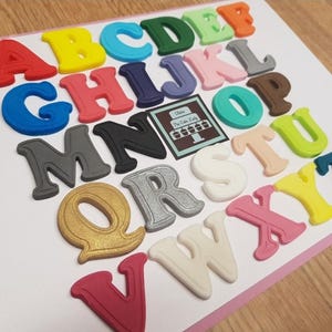 May include: A selection of brightly coloured alphabet letters in shades like red, yellow, and blue. The letters are displayed on a white surface, with a small card that says "The Cake Lady".