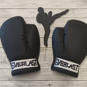 May include: Two black boxing gloves with white "EVERLAST" lettering on the bottom of each glove. The gloves are on a wooden surface.