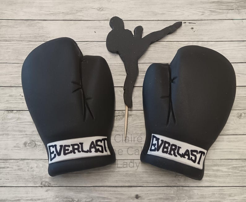 May include: Two black boxing gloves with white "EVERLAST" lettering on the bottom of each glove. The gloves are on a wooden surface.