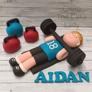 May include: A cake decoration showing a figure lifting a barbell, wearing a blue shirt with the number 18. Red and blue kettlebells are nearby. The name "AIDAN" is spelled out in blue letters. The cake is made by "Claire The Cake Lady".
