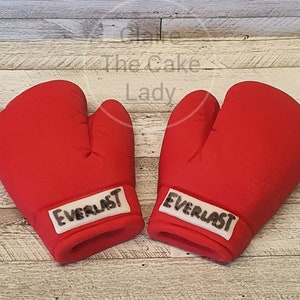 May include: Two red boxing gloves with white Everlast branding. The gloves are made of fondant and are perfect for a boxing themed cake.