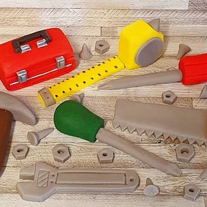 May include: A set of red, yellow, green and brown toy tools, including a hammer, saw, screwdriver, wrench, tape measure and toolbox. The tools are surrounded by small grey and brown plastic nuts and bolts.