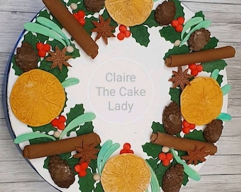 May include: A white cake decorated with a Christmas wreath made of green leaves, orange slices, cinnamon sticks, star anise, pine cones, and red berries. The cake is decorated with the text "Claire The Cake Lady".