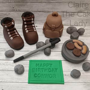 May include: A green cake topper with the text "Happy Birthday Connor" and a camping theme. The topper includes a brown backpack, two brown hiking boots, a grey hiking stick, a grey campfire with sausages and a few grey rocks.