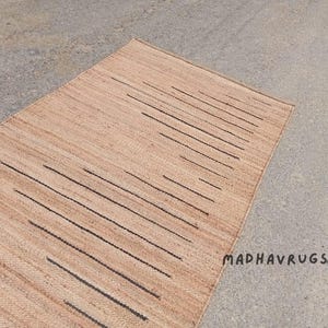 May include: A natural fiber rug with a geometric pattern of black lines on a light brown background. The rug is made of jute and is perfect for adding a touch of natural beauty to any room. MADHAVRUGS