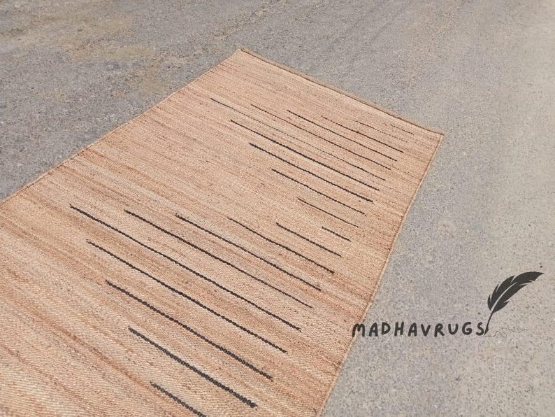 May include: A natural fiber rug with a geometric pattern of black lines on a light brown background. The rug is made of jute and is perfect for adding a touch of natural beauty to any room. MADHAVRUGS