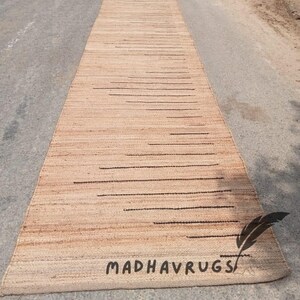May include: A long, narrow, natural fiber rug with a subtle striped pattern and black lines. The rug is made of jute and has a natural, earthy color. The rug is laid out on a gray asphalt surface. The text "MADHAVRUGS" is visible in the lower right corner of the rug.
