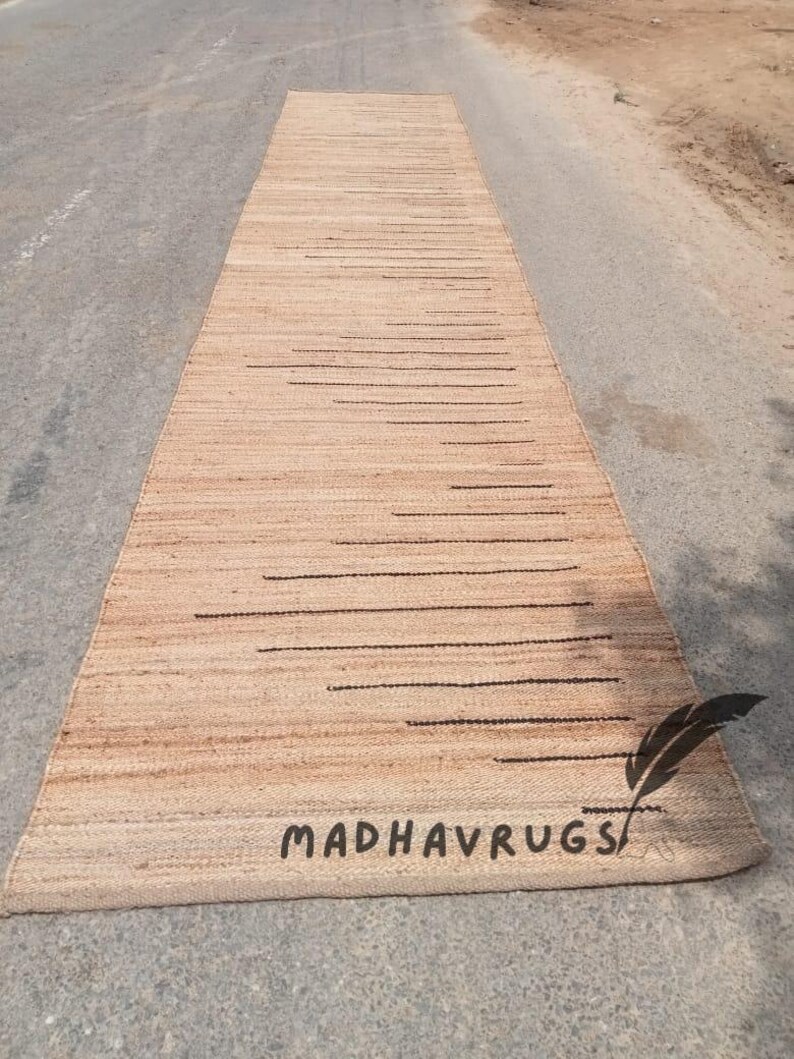 May include: A long, narrow, natural fiber rug with a subtle striped pattern and black lines. The rug is made of jute and has a natural, earthy color. The rug is laid out on a gray asphalt surface. The text "MADHAVRUGS" is visible in the lower right corner of the rug.