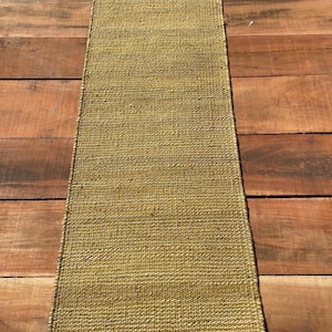 May include: A rectangular, woven rug in shades of olive green and tan. The rug has a textured surface and a simple, minimalist design. The rug is placed on a wooden surface.
