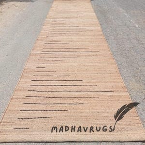 May include: A long, natural fiber rug with a striped pattern in a light brown color. The rug is rolled up and has the text "MADHAVRUGS" printed on it.