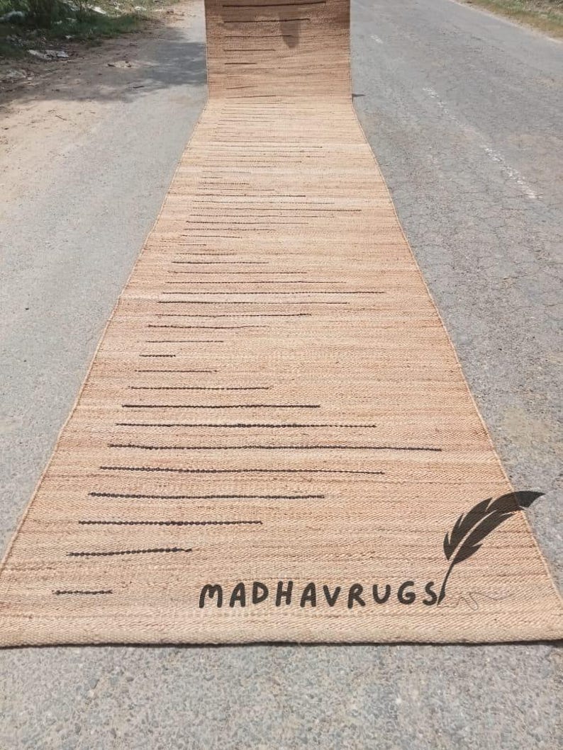 May include: A long, natural fiber rug with a striped pattern in a light brown color. The rug is rolled up and has the text "MADHAVRUGS" printed on it.