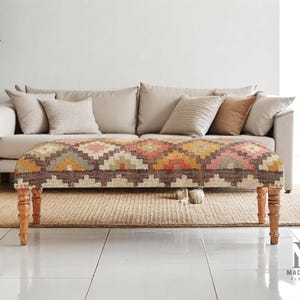 Pode incluir: Banqueta retangular com um padrão geométrico colorido em tons de castanho, rosa, laranja e verde. A banqueta tem pernas de madeira e está colocada em frente a um sofá bege. A sala tem uma paleta de cores neutras.