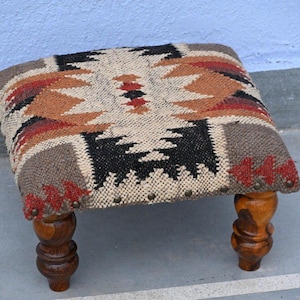 Può includere: Uno sgabello quadrato con una parte superiore in tessuto intrecciato con un motivo geometrico nei toni del crema, marrone, nero e rosso. Lo sgabello è sostenuto da quattro gambe in legno tornito con finitura lucida e borchie metalliche.