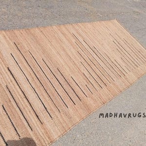 May include: A natural fiber rug with a woven pattern of vertical black lines on a light brown background. The rug is made of jute and is perfect for adding a touch of natural beauty to any room.