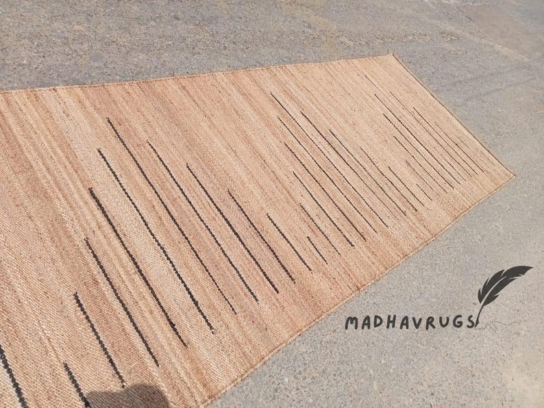 May include: A natural fiber rug with a woven pattern of vertical black lines on a light brown background. The rug is made of jute and is perfect for adding a touch of natural beauty to any room.