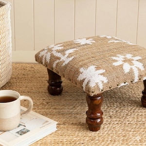 May include: A square footstool featuring a natural tan jute top with a white floral pattern and brass nailhead trim. The stool is supported by four dark brown turned wooden legs, providing a sturdy base for this low-profile furniture piece.