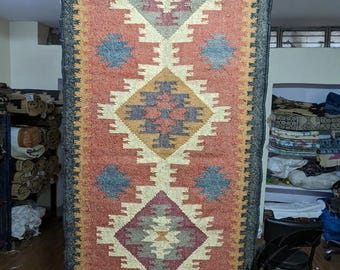 May include: A long, narrow rug with a geometric pattern in shades of red, blue, beige, and brown. The rug has a fringe border and is made of natural fibers.