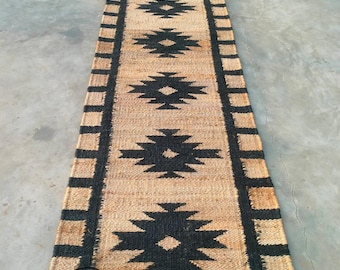 May include: A long, narrow rug with a black and tan geometric pattern. The rug has a black border with a repeating pattern of stripes. The rug is made of natural fibers and has a rustic look.