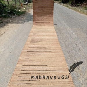 May include: A long, narrow, natural fiber rug with a striped pattern in a light brown color. The rug has black lines running horizontally across the surface. The rug is rolled up and lying on a paved road. The text "MADHAVRUGS" is visible on the rug.