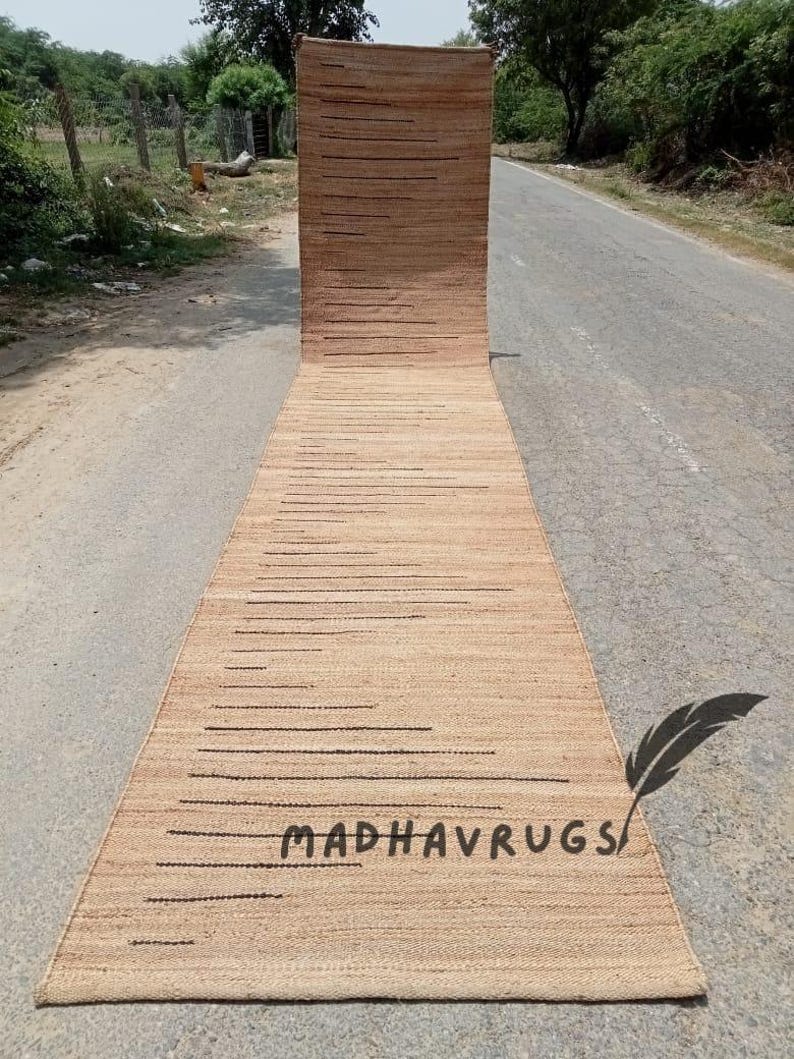 May include: A long, narrow, natural fiber rug with a striped pattern in a light brown color. The rug has black lines running horizontally across the surface. The rug is rolled up and lying on a paved road. The text "MADHAVRUGS" is visible on the rug.