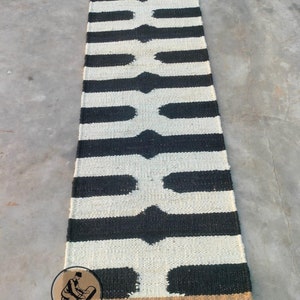 Puede incluir: Alfombra rayada en blanco y negro con un patrón geométrico. La alfombra está hecha de fibras naturales y tiene un borde de yute.