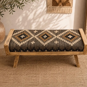 May include: A wooden bench with a patterned cushion. The cushion features a geometric design in shades of gray, black, and tan. The bench has a natural wood frame and is placed on a woven rug. The bench is a piece of furniture.