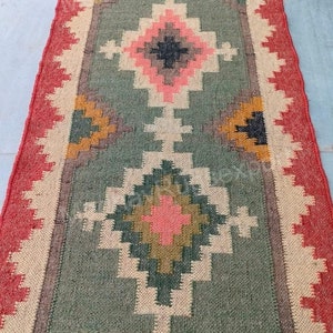 May include: A green, red, and beige patterned rug with a geometric design. The rug has a fringe border.